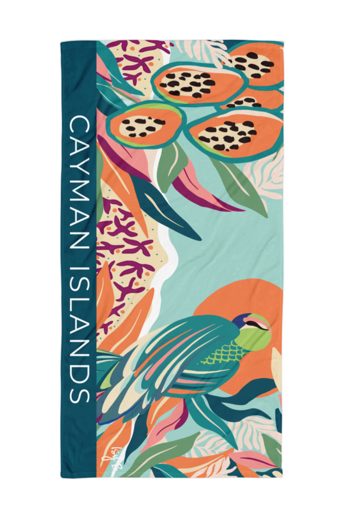 A rectangular beach towel featuring a vibrant tropical print of a parrot, papayas, and coral, with a dark teal vertical band on the left side printed with the text 'CAYMAN ISLANDS' in white.