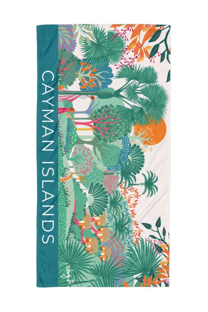 A rectangular beach towel featuring a colorful tropical landscape print with palm trees, houses, and a teal vertical band on the left side that reads 'CAYMAN ISLANDS' in white text.