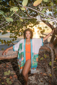 Woman in the Sundowner orange Hero swimsuit and Kaiso Kimono with Fringe standing under a tree with a sunset over water in the background.