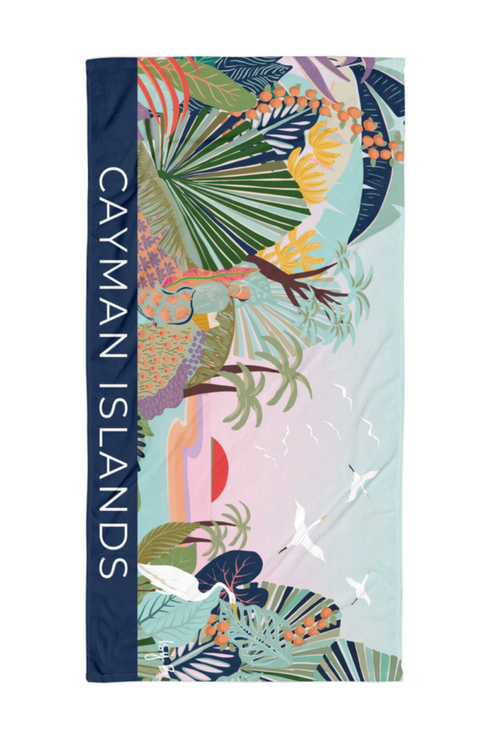 A beach towel featuring a colorful tropical print with palm leaves, flamingos, birds, and a pastel landscape, bordered by a navy blue side panel with the text 'CAYMAN ISLANDS' in white.