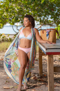 Woman in a bikini standing by a wooden table with a Lekki Silk Modal Pareo Sarong draped over it on a beach.