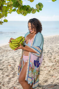 Woman holding a bunch of bananas on a beach with ocean and sky in the background wearing the Lekki Wrap Kimono with Belt