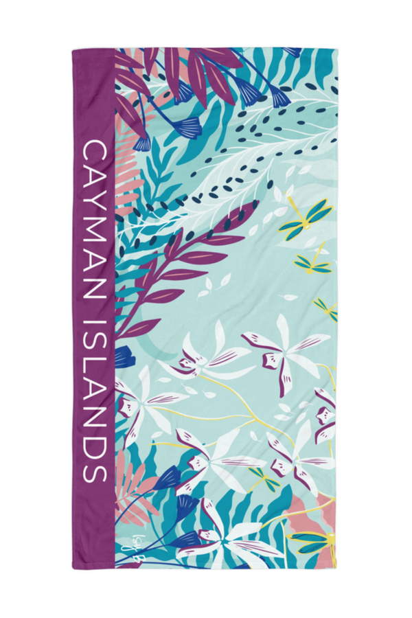 A rectangular beach towel featuring a tropical floral print with the words 'CAYMAN ISLANDS' printed vertically in white on a purple side panel, set against a plain white background.