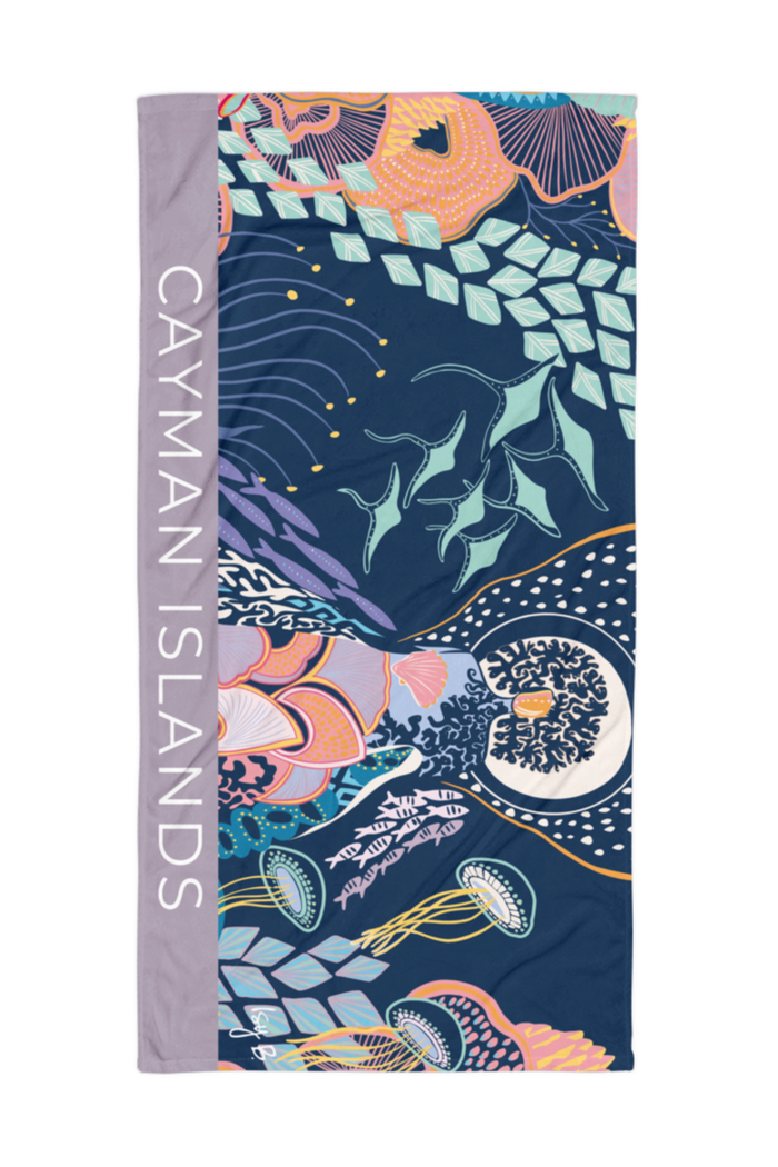 A rectangular beach towel featuring a colorful underwater design with sea creatures on a dark navy background, with a lavender-gray border on the left side printed with the text 'CAYMAN ISLANDS' in white.