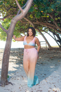 Woman in a colorful bikini and Sunset Beach Lounge Pants standing on a sandy beach with trees in the background